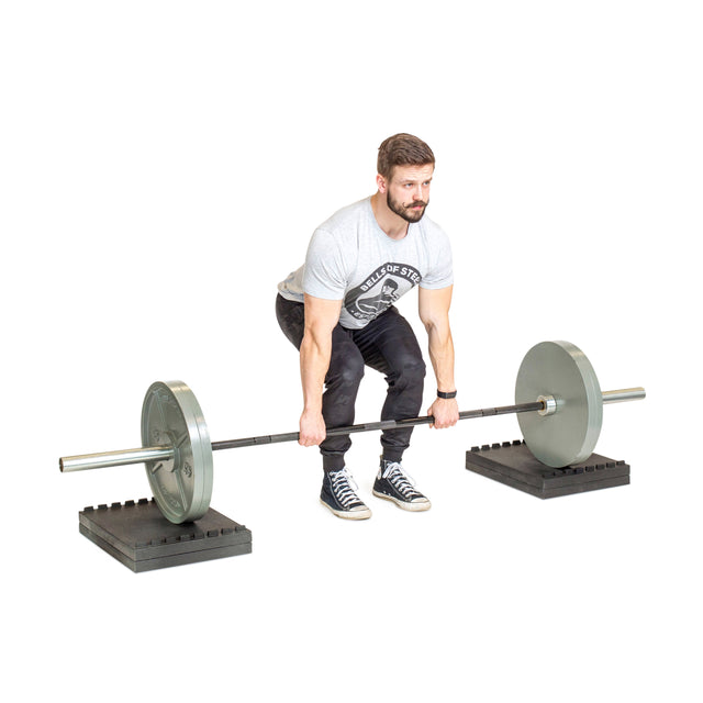 A man in a gray T-shirt and black pants prepares to deadlift a barbell resting on Bells of Steel Stackable Pull Blocks. He grips the bar with both hands, ready to lift, against a plain white background.