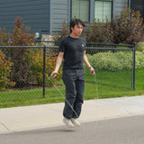 Wearing black athletic clothing, a person uses the Bells of Steel Ultimate Backyard Training Bundle to jump rope with fitness equipment on a sidewalk in front of a house with a chain-link fence and green bushes, enjoying an outdoor workout experience.