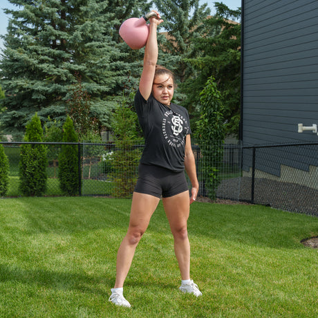 A woman stands on grass, holding a pink Bells of Steel Competition Kettlebell overhead with one arm. She wears a black t-shirt, black shorts, and white shoes. Trees and a fence are visible in the background.
