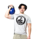 A person in a grey Bells of Steel t-shirt holds a blue Bells of Steel Competition Kettlebell on their shoulder, standing against a plain white background.