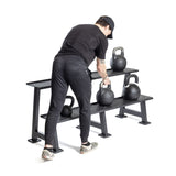 A person in black workout gear and checkered sneakers picks up a competition kettlebell from the Bells of Steel Kettlebell Rack, which holds multiple kettlebells—ideal inspiration for your home gym, all set against a plain white background.