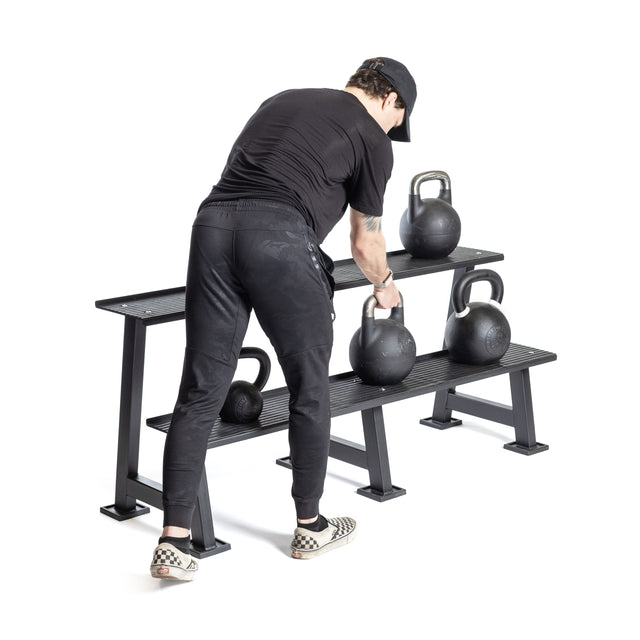 A person in black workout gear and checkered sneakers picks up a competition kettlebell from the Bells of Steel Kettlebell Rack, which holds multiple kettlebells—ideal inspiration for your home gym, all set against a plain white background.