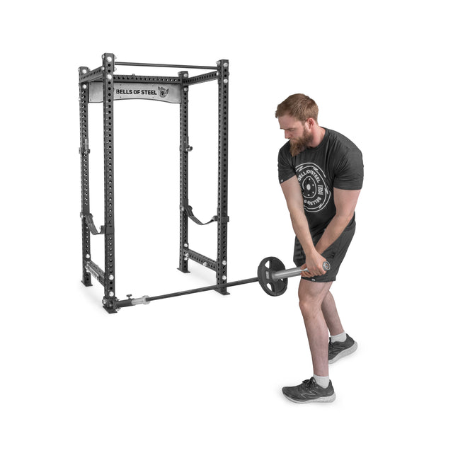 A man in athletic wear performs a strength workout, lifting a barbell from the UHMW-lined Landmine Rack Attachment by Bells of Steel, using both hands with unwavering focus.