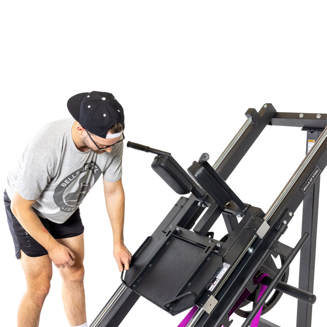 A man in a gray t-shirt, black shorts, and a backward cap adjusts the Bells of Steel Leg Press Hack Squat Machine with metal rails, showcasing versatile home gym equipment against a white background.