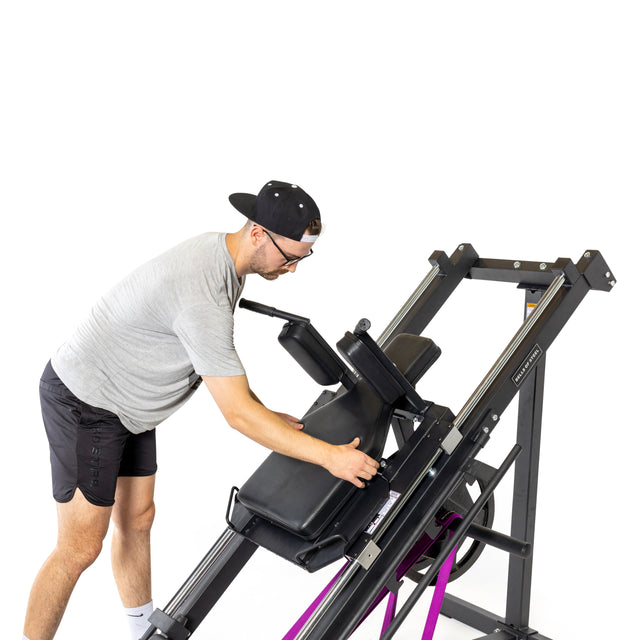 A man in a gray shirt and black shorts adjusts the seat of a Bells of Steel Leg Press Hack Squat Machine with resistance bands, ideal for home gyms, set against a white background.