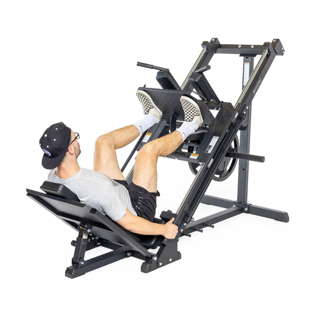A man in sportswear and a cap uses the Bells of Steel Leg Press Hack Squat Machine, pushing against the footplate with both legs while lying on the padded bench. Side view on a white background.