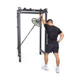 A man in black athletic wear performs a landmine press with a barbell and weight plate in his home gym, standing in front of the Bells of Steel Manticore Four Post Power Rack - Prebuilt (3" x 3", 1" Holes).