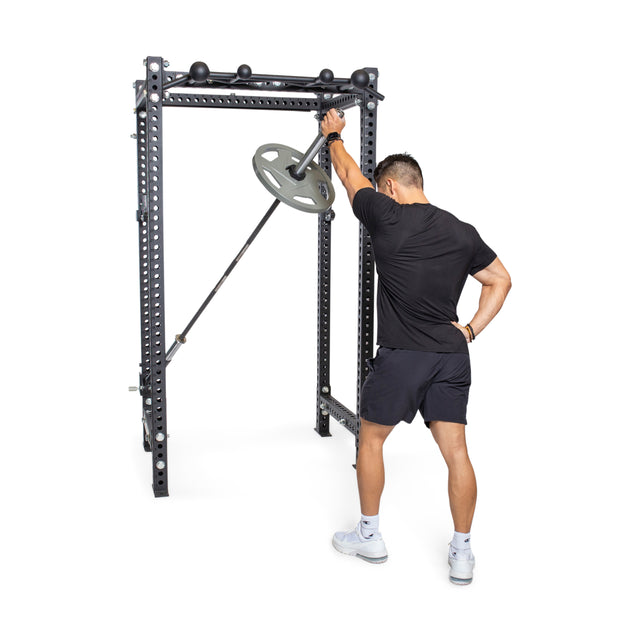 A person dressed in a black shirt and shorts works out using a landmine attachment on the Bells of Steel Manticore Four Post Power Rack - Prebuilt (3" x 3", 1" Holes). They are positioned to press a barbell with a weight plate, utilizing its versatile design that is ideal for home gyms.