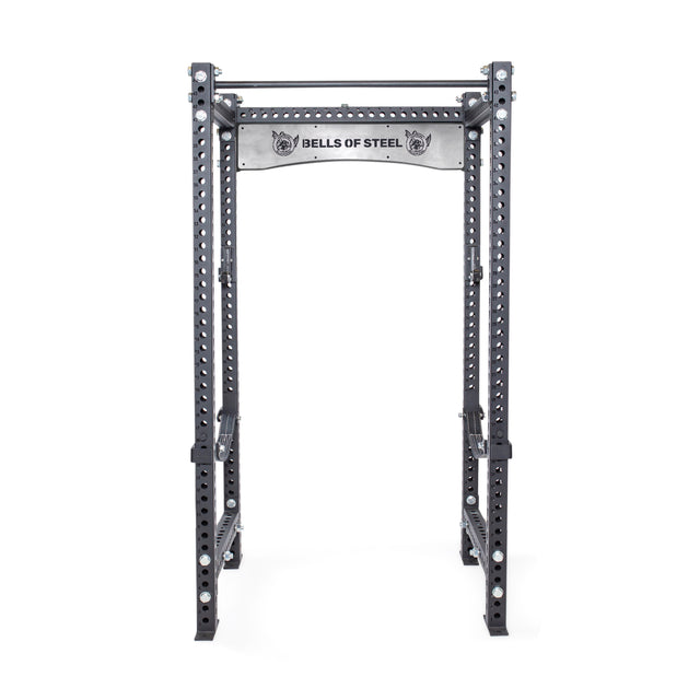 The Manticore Four Post Power Rack - Prebuilt by Bells of Steel, crafted from black steel, is perfect for home gyms. It boasts a modular design with a pull-up bar and features 1" holes for adjustable attachments. The top includes a branded plate displaying the "Bells of Steel" logo.
