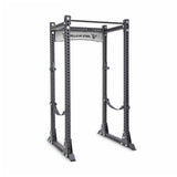 The Manticore Flat Foot Power Rack by Bells of Steel is a sturdy, modular unit with adjustable pegs and safety bars, featuring a pull-up bar. Its black metal frame displays the "Bells of Steel" branding on a horizontal support beam.