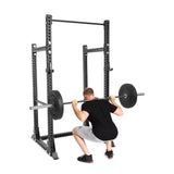 A man performs a barbell squat in his home gym using the Bells of Steel Manticore Half Rack - Prebuilt (3" x 3", 1" Holes), holding the weighted barbell across his shoulders.