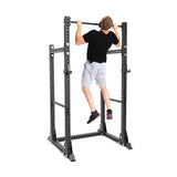 A person in a black shirt and gray shorts is doing a pull-up on the Bells of Steel Manticore Half Rack - Prebuilt (3" x 3", 1" Holes), perfect for any home gym, against a white background.