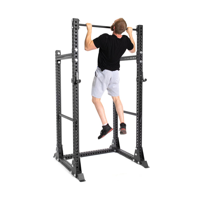 A person in a black shirt and gray shorts is doing a pull-up on the Bells of Steel Manticore Half Rack - Prebuilt (3" x 3", 1" Holes), perfect for any home gym, against a white background.