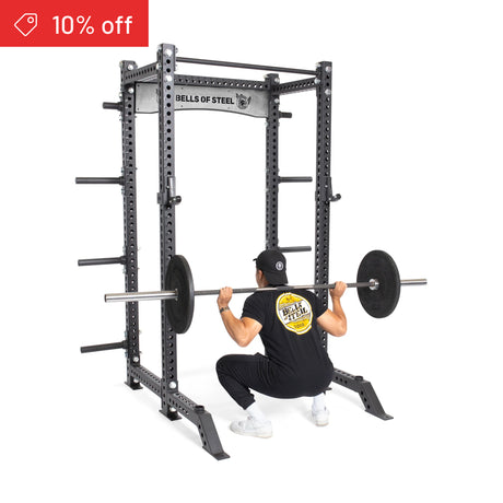 A person in a black shirt and cap performs a barbell squat in the Bells of Steel Manticore Collegiate Power Rack Builder (3" x 3", 1" holes), with weight plates loaded. A "10% off" sale tag appears in the top-left, highlighting this sturdy rack.