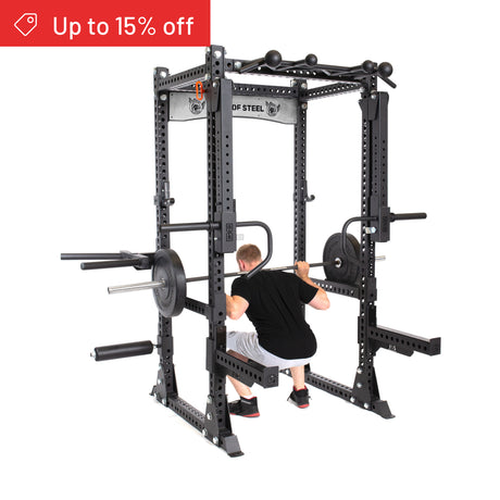 A man squats with a barbell inside the Bells of Steel Manticore Flat Foot Power Rack Builder (3" x 3", 1" Holes), which has various attachments and holds weight plates. A red "Up to 15% off" banner appears in the top left corner.