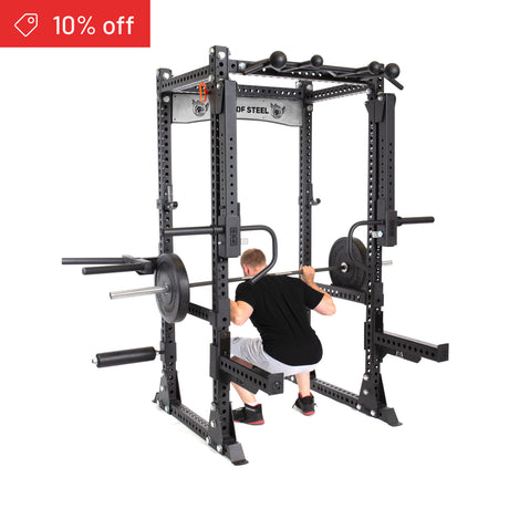 A man squats with a barbell inside the Bells of Steel Manticore Flat Foot Power Rack Builder (3" x 3", 1" holes), loaded with weight plates. A red "10% off" tag appears in the top left for your home gym upgrade.
