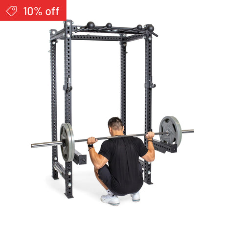 A man squats with a Manticore Four Post Power Rack Builder (3" x 3", 1" Holes) by Bells of Steel. A red "10% off" banner appears in the top left—ideal for upgrading your home gym. White background, 3x3 uprights inside a power rack.