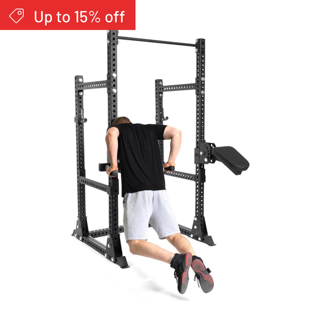 A person in a black shirt and gray shorts performs dips on the Bells of Steel Manticore Half Rack Builder (3" x 3", 1" Holes). A red banner in the top left reads "Up to 15% off." Perfect for any home gym.