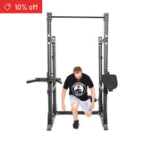 A man in a black t-shirt and white shorts kneels inside the Bells of Steel Manticore Half Rack Builder (3" x 3", 1" holes), ideal for home gyms. A red "10% off" banner is shown in the top left, with a plain white background.