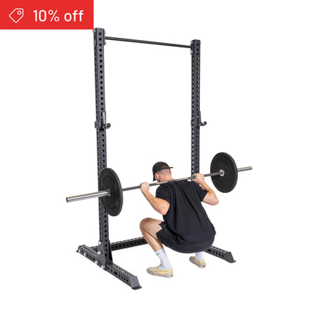 A person in athletic wear squats with a barbell on the Bells of Steel Manticore Squat Stand Builder (3" x 3", 1" Holes), under a black rack. "10% off" appears in the top left. White background, ideal for home gyms.