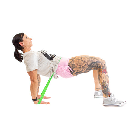 A woman performs a hip thrust using Bells of Steel Mini Bands to increase intensity, supporting herself with her hands and feet on the ground, hips raised, against a white background. Tattoos are visible on her legs.