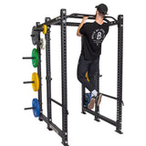 A man in a black shirt and cap does a pull-up on the Bells of Steel Multi-Grip Pull Up Bar attached to a black power rack with colorful weight plates stored on the side, highlighting his strength training routine.