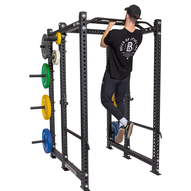 A man in a black shirt and cap does a pull-up on the Bells of Steel Multi-Grip Pull Up Bar attached to a black power rack with colorful weight plates stored on the side, highlighting his strength training routine.