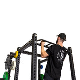 A man in a black T-shirt and cap performs a pull-up on the Bells of Steel Multi-Grip Pull Up Bar, which is mounted on a black power rack with weight plates, showcasing strength training against a white background.
