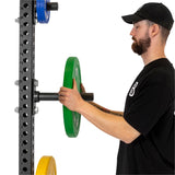 A person in a black shirt is adding an Olympic-sized weight plate onto a horizontal bar of the weight rack, which is equipped with Bells of Steel Bolt-On Plate Peg Sets and features several plates in green, blue, and yellow hues organized at different levels for optimal storage.