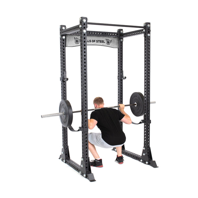 Inside the Manticore Flat Foot Power Rack by Bells of Steel, a person performs a squat with a barbell. The rack supports up to 1,000 pounds and displays the brand's label. The individual wears a black shirt and gray shorts.