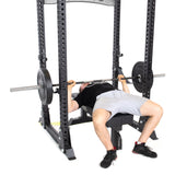 The individual lies on the Manticore Flat Foot Power Rack by Bells of Steel, lifting a barbell with weights and modular components. The setup supports up to 1,000 pounds. They're dressed in a black shirt, gray shorts, and red-accented shoes against a plain white background.