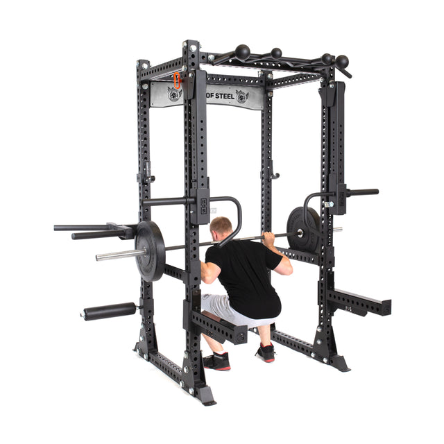A man in a black shirt and shorts squats confidently with a barbell inside the sturdy Manticore Flat Foot Power Rack by Bells of Steel, which boasts a 1,000-pound capacity and features attachments like weight storage pegs, safety bars, and pull-up handles.