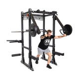 A man in a black t-shirt and gray shorts does shoulder presses with the sturdy Manticore Flat Foot Power Rack - Prebuilt by Bells of Steel. This black equipment, featuring modular components and attached weights, boasts 3" x 3" dimensions and convenient 1" holes for attachments.