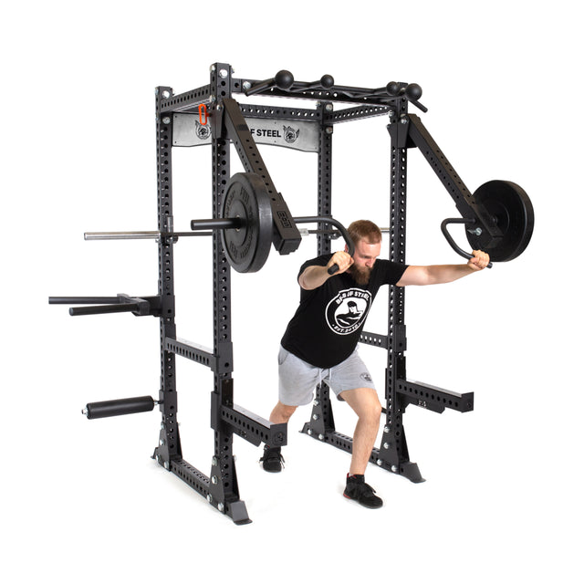A man in a black t-shirt and gray shorts does shoulder presses with the sturdy Manticore Flat Foot Power Rack - Prebuilt by Bells of Steel. This black equipment, featuring modular components and attached weights, boasts 3" x 3" dimensions and convenient 1" holes for attachments.