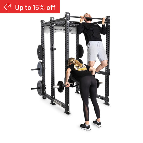 A woman lifts a barbell while a man does pull-ups on the Bells of Steel Manticore Six Post Power Rack Builder (3"x3", 1" holes) with weight plates. A red banner in the top left corner reads "Up to 15% off".