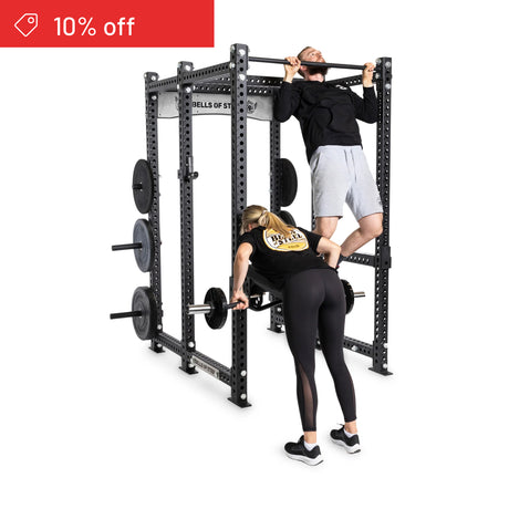A man does pull-ups on the Bells of Steel Manticore Six Post Power Rack Builder (3" x 3", 1" Holes), while a woman lifts a barbell below. Weight plates are stored on the rack, with a red "10% off" label in the top left corner.