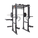 A Bells of Steel Manticore Flat Foot Power Rack in black set up for squats with a barbell and weights. It features pull-up bars, safety bars, modular components with various attachments, and a 1,000-pound capacity, with the brand logo visible on the top crossbar.
