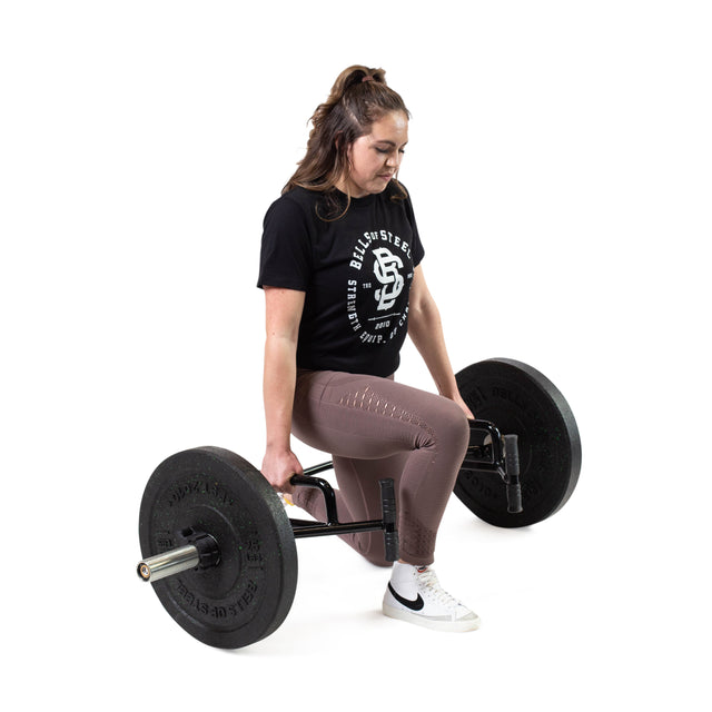 A woman in a black t-shirt, mauve leggings, and white sneakers performs a lunge with the Bells of Steel Open Trap Bar / Hex Bar against a white background.