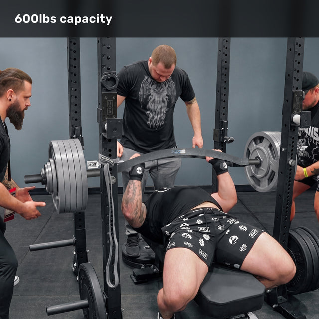 A man on a weight bench gets ready to lift a heavily loaded Bells of Steel Arch Nemesis Swiss Bar, assisted by three spotters. Text above states "600lbs capacity.