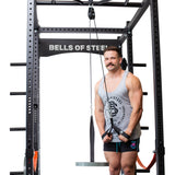 A muscular man in a grey tank top and black shorts does tricep pushdowns on the Bells of Steel Cable Pulley, gripping the handle with both hands and smiling at the camera.