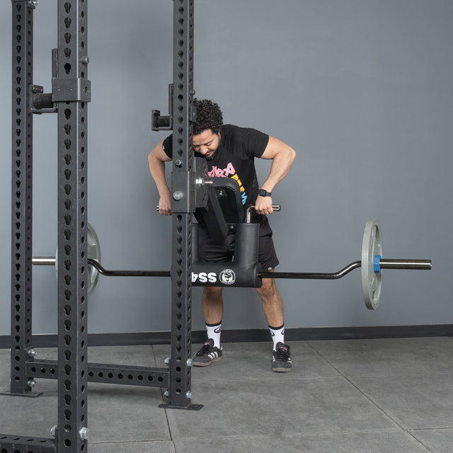 Wearing athletic clothes, a person performs a chest-supported row with the Bells of Steel Safety Squat Bar - The SS4 attached to a squat rack in a gym featuring gray walls and a tile floor.