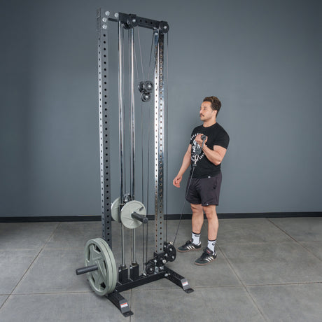 A man stands by a tall, black cable machine in a gym with gray walls and a concrete floor, using Bells of Steel Fat Bar - Single D Handle attachments to enhance grip strength.