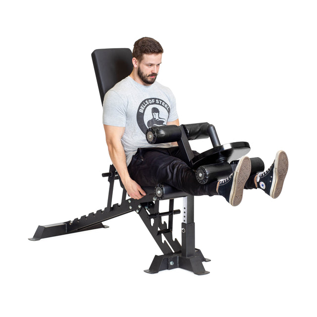 A man in a gray T-shirt and black pants uses a leg extension machine while seated on the Bells of Steel Buzz-Saw Heavy-Duty Adjustable Bench, which features premium vinyl upholstery.