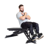 A bearded man sits on the Bells of Steel Buzz-Saw Heavy-Duty Adjustable Bench, arms crossed and feet secured under padded rollers, performing a fitness exercise against a plain white background.