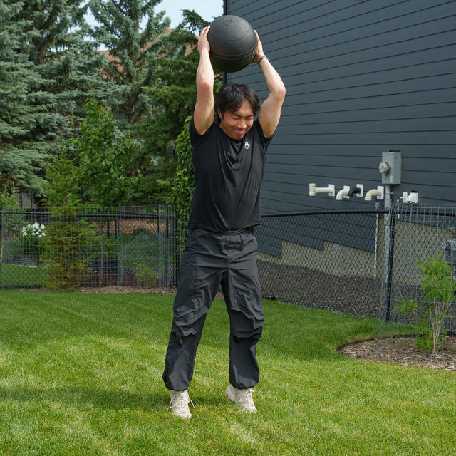 Wearing black clothing, a person stands on the grass of a backyard gym, lifting a medicine ball overhead with both hands as they prepare to slam it down as part of an outdoor workout using the Bells of Steel Ultimate Backyard Training Bundle.