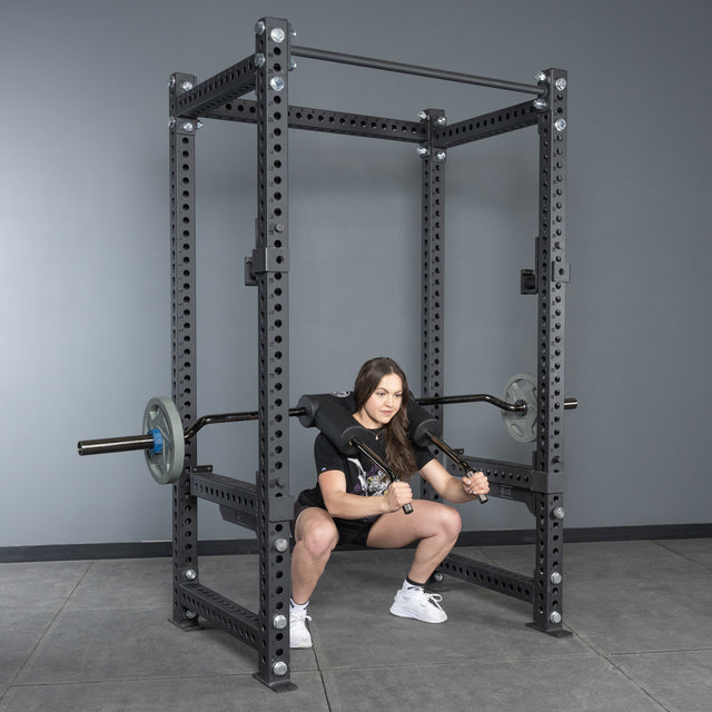 A woman does squats in a gym power rack using the Bells of Steel Safety Squat Bar - The SS4, with weight plates on her shoulders. She wears a black T-shirt, black shorts, and white sneakers.