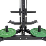 A close-up of the All-in-One Trainer features green 10 kg weight plates labeled "Bells of Steel." The image highlights the sturdy base and squat stand-like support, emphasizing its durability and reliability in strength training.