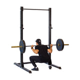 A person in black athletic wear and a cap is performing a barbell squat using the Bells of Steel Hydra Squat Stand - Prebuilt (3" x 3", ⅝" Holes), with the barbell resting across their shoulders.