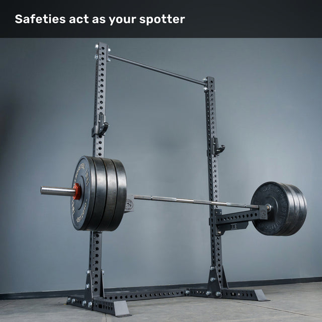 A barbell loaded with heavy weight plates rests on the safeties of the Bells of Steel Manticore Squat Stand - Prebuilt (3" x 3", 1" Holes), featuring robust black uprights. White text above reads, "Safeties act as your spotter." The minimalist scene, set against a plain gray wall and floor, is ideal for any home gym.