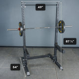 The Bells of Steel Manticore Squat Stand - Prebuilt (3" x 3", 1" Holes), a black metal rack with sturdy 3x3 uprights, is pictured complete with a barbell and weights. Measuring 87 1/4" tall, 49" wide, and 54" deep, this squat stand is perfect for any home gym. The floor and wall are gray.